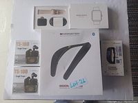 Photo showing two boxed TS-100 wireless earbuds, two boxed smart watches including a rose gold watch and a Peant branded fitness tracker, and a boxed Monster Boomerang Bluetooth personal wearable speaker arranged on a white background.