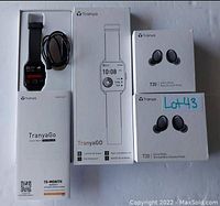 Photo showing TranyaGo Smartwatch with charging cable and user manual alongside two boxes of Tranya T20 wireless earbuds, one for game mode bass boosted, the other with normal/bass boosted modes.