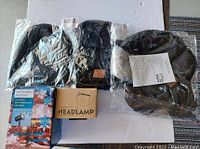 Photo showing three black Bluetooth beanies in plastic packaging, snowflake projection light box, and black headlamp with packaging, all arranged on a white surface.