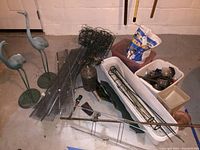 Two metal heron statues, white plastic planter pots, metal mesh pieces, solar lights, garden fencing, ice melt bag, copper pipe, hand seed spreader, green glass birdbath base, and a lantern-shaped metal solar light holder arranged on the basement floor.