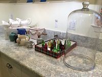 Photo showing the full lot on a kitchen counter including colored glass bottles, white ceramic pitchers, pewter cup, glass stein, and the large clear glass dome with a crack.