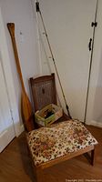 Wood Beaver paddle, fishing rod with reel leaning against wall, folding cane chair, and upholstered footstool on floor.