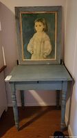 Blue rustic flip top wooden desk with turned legs positioned against wall, with framed child portrait art leaning on desk.