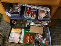 Shelving unit with several plastic bins containing dolls and accessories, plus dollhouse-related books and boxed items arranged on shelves and floor space.