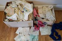 Two boxes filled with various vintage linens including embroidered handkerchiefs, napkins, tablecloths, doilies, and handmade potholders; some pieces draped outside on wood floor.