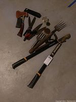 Various hand tools laid out on concrete floor including Estwing hatchet with leather sheath, sharpening stone, Fiskars grass clippers, red-handled clipping shears, metal garden hand tools, flower frogs, and hose nozzles.