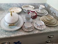 Overview photo showing the assortment of fine china on a countertop including lidded serving dish, plates, bowls, and teacups from various brands.