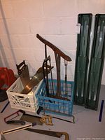 Photo showing wooden-handled saws including a large two-person saw, smaller hand saws, and small folding saws arranged in plastic crates alongside saw horses.
