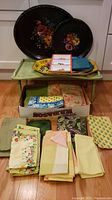 Photo displaying wooden lap tray table, two round floral trays, and assorted rectangular trays on top with boxed linens below on wooden floor.