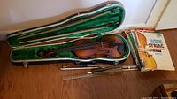 Overview of violin in case with two bows alongside a stack of instruction and kid singing books on the floor.