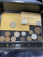 Coins arranged in a black tray, including a 1961 penny with a hole, multiple old-world coins, and a Littleton Stamp and Coin Company card showing Brazil coin set.