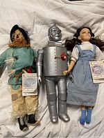 Three Wizard of Oz dolls arranged side by side on white fabric background showing full front view of Scarecrow, Tin Man, and Dorothy dolls with tags attached.