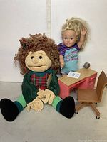 Folkmanis girl puppet sitting next to blonde-haired doll on a small desk with rolling chair nearby