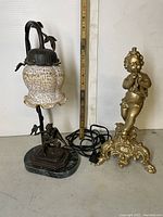 Both lamps together showing size relative to measuring stick; marble based wrought iron lamp with white textured glass shade and metal cherub lamp base with ornate design