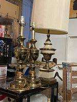Three vintage brass lamps grouped on a glass table; two are matching brass urn-shaped lamps and one is a ceramic and brass lamp with a fabric shade.