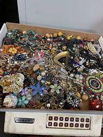 Top-down view of a box filled with mixed broken jewelry, beads, and findings including colorful floral brooches, chains, and beads in varied colors and materials.