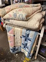 Four folded vintage quilts displayed on a white painted wooden quilt holder in a cluttered room area. Quilts vary in color and pattern, with visible rings on the large arch quilt for mounting.
