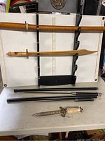 Two wooden practice swords mounted on a black wooden wall rack with curved slots, three black segmented rods, and one decorative dagger with ornate handle and etched blade lying on a white table.