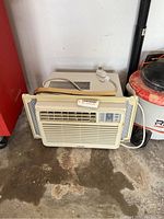 Front view of beige Samsung window air conditioner with control panel, remote on top, three-prong power cord, and adjustable accordion side panels, sitting on concrete floor.