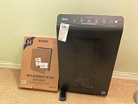 Winix air purifier next to boxed replacement filter and remote control on floor.