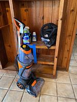 Front and side views of Bissell ProHeat Pet Revolution upright vacuum with cleaning bottles and accessories on a wooden shelf and tiled floor.