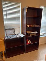 Two wooden bookcases placed next to each other; the taller one with five shelves contains some items, the shorter has two shelves and some boxed items displayed on top.