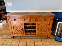 Front view of oak cabinet with two drawers, two paneled doors with X-shaped design, central wine rack holding nine bottles, and slate tile top.