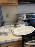 Four Corning Ware dishes with lids, one casserole dish and one platter on a kitchen counter near a stove