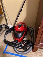 Red Dirt Devil bagless vacuum cleaner with hose, wand, and attachments positioned on carpet near wooden furniture.