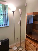 Two floor lamps standing side by side against a pale green wall next to a window and cabinet.