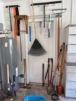 Photo showing garden tools including leaf rake hanging on wall, shovel, soil cultivator, bow rake and hedge loppers placed near door