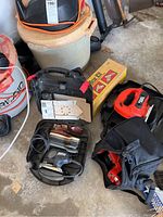 Black & Decker jigsaw in black hard case, surrounded by other items including an orange and black tool, a yellow box, and sandpaper sheets.