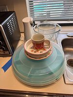 View of the lot including large blue plastic carousel, child's dish set with cup and small dish, Oster can opener, and clear glass mixing bowls on kitchen counter