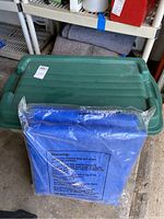 Green plastic storage bin with lid and folded grey moving blankets on shelf in background