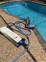 Overview of Zodiac MX6 pool cleaner with attached hoses coiled poolside and cardboard box containing extension hoses.