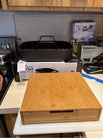 Black nonstick All-Clad roaster with rack on box, wooden K-Cup holder, and wooden cutting board on counter.