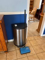 Aluminum trash can with foot pedal and Bissell sweeper leaning against the wall on tiled floor.