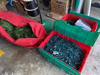 Red bag with artificial tree branches next to large plastic bin containing tangled strings of Christmas lights and cords, with visible green metal tree stand base.