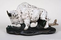 White ceramic buffalo figurine showing front-left side, standing on black textured base with branch detail and chipped horn.