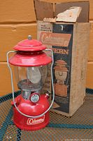 Red Coleman lantern standing in front of its original cardboard box, on a metal table