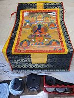 Full view of silk Thangka artwork folded on floor showing multi-patterned borders and central painted panel with multiple Buddha figures.
