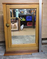 Front view of the vintage wooden medicine cabinet with beveled mirror door, showing the reflective mirror surface and brass latch, ruler indicating size next to cabinet.