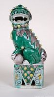 Front-side view of the green porcelain Foo Dog showing detailed facial features, open mouth and ornamentation with yellow nodules and purple accents on the head and body, sitting on a decorated square pedestal base.