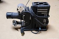 Side view of Tri-Purpose Projector Model AAA showing lens, power cord, and film reel spindles on black metal body