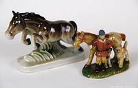 Photo showing two ceramic figurines, a dark brown horse jumping over water, and a tan horse with boy and dogs