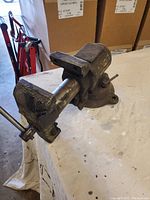 Front and side view of a black metal bench vise mounted on a workbench, showing jaws and base with swivel mechanism.