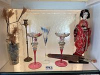 Photo showing a 17-inch Asian doll dressed in a red kimono on a small stand, two pink-stemmed glasses with colorful bowls, one tall clear glass with blue design, two brass candlesticks (one damaged), an Asian-style folding umbrella with wooden handle, creamer, and dried floral stems in a cut glass vase.