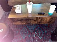 Front view of rectangular wood coffee table with hard clear plastic top covering. Shows items placed on top including tissue box, large cup with straw, remote, and white-blue cylindrical object.