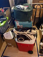 View of stacked items including red wheeled cooler at bottom, vintage camera with white and black bag on top, dark green Crock-Pot carrying case on top, and Nostalgia ice cream maker box beneath cooler handle.