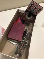 Photo of open shoebox with assorted cosmetic items including a pink cosmetic bag, lip gloss, and small bottle with pump
